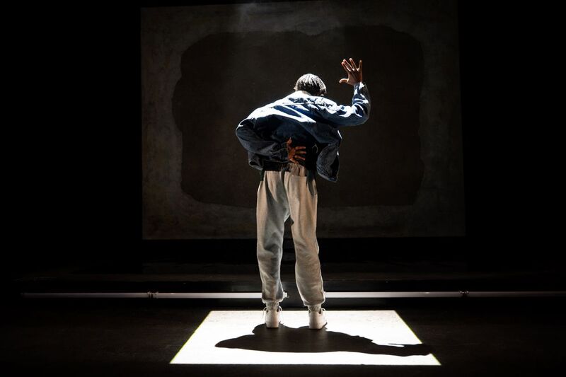 Picture of Selorm Adonu on stage facing away from the camera. One hand is held behind his back and the other is held up.