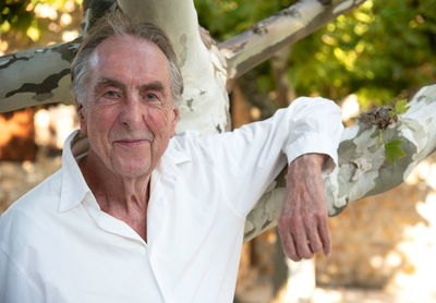 Eric Idle wearing a white shirt leaning on a tree branch