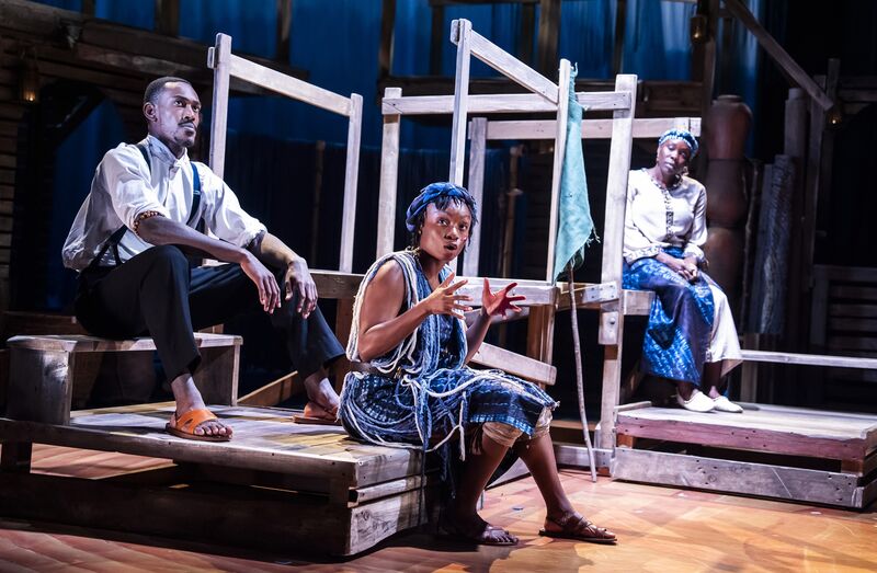 A black woman in an indigo outfit sits on top of a wooden crate, sat behind her, slightly hiigher up is a black man with his arms resting on his knees. To their left is another black woman looking toward them, she is also sat on a wooden structure.