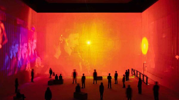 A large immersive exhibition space bathed in vivid red-orange light, with a glowing geometric cross-like pattern projected on the far wall and visitors silhouetted throughout the room.