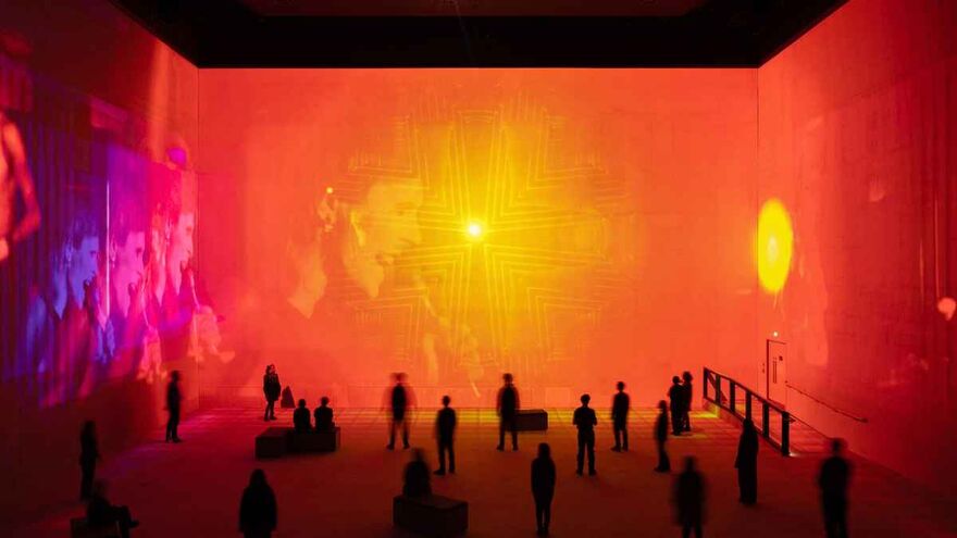 A large immersive exhibition space bathed in vivid red-orange light, with a glowing geometric cross-like pattern projected on the far wall and visitors silhouetted throughout the room.