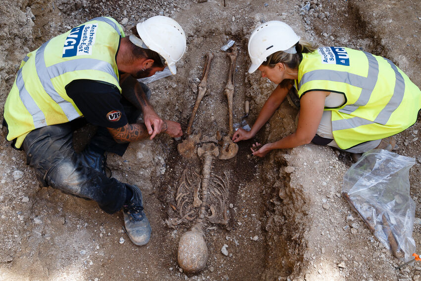 Two people in high vis jackets and white hard hats are crouched over an uncovered skeleton of a person
