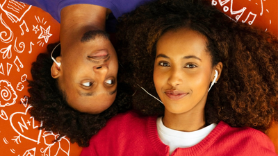 Two people with curly hair lie head-to-head on a bright orange background, sharing white wired earbuds. One person, wearing a purple shirt, looks sideways at the other, who wears a red sweater over a white shirt and smiles softly at the camera. White doodles of musical notes, stars, hearts, and swirls surround them, creating a playful, music-themed scene.