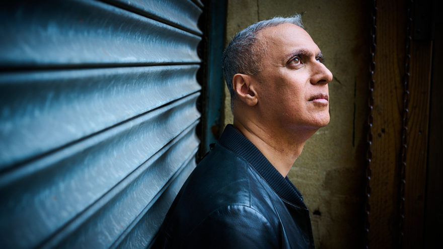 Nitin Sawhney stood against metal shutters looking dramatically away from camera