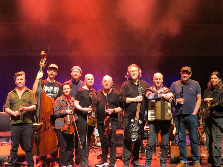 9 people standing on a stage with a red smokey background. Each people is holding an instrument, including a double bass, guitar, violins and an accordian