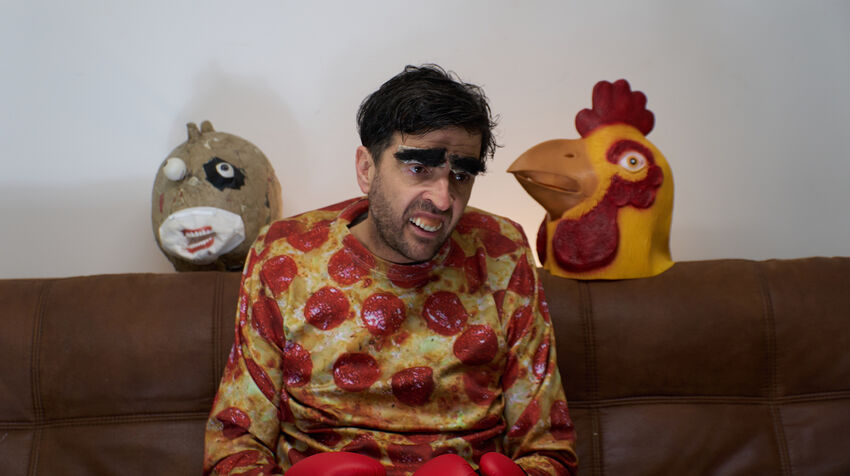 Spencer Jones sat on a sofa wearing a t-shirt that looks like a pepperoni pizza, with a large model of a bird over each shoulder