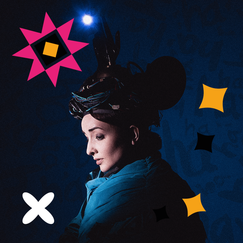 Julia Masli standing dramatically in a blue coat and hat, pops of bright colour burst from the dark background