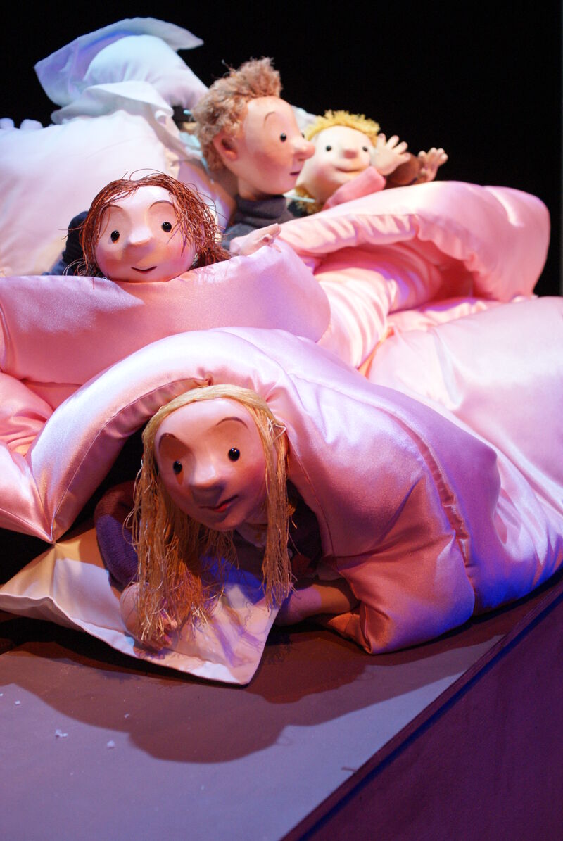 Three wooden puppets of children bundled up in a satin pink duvet