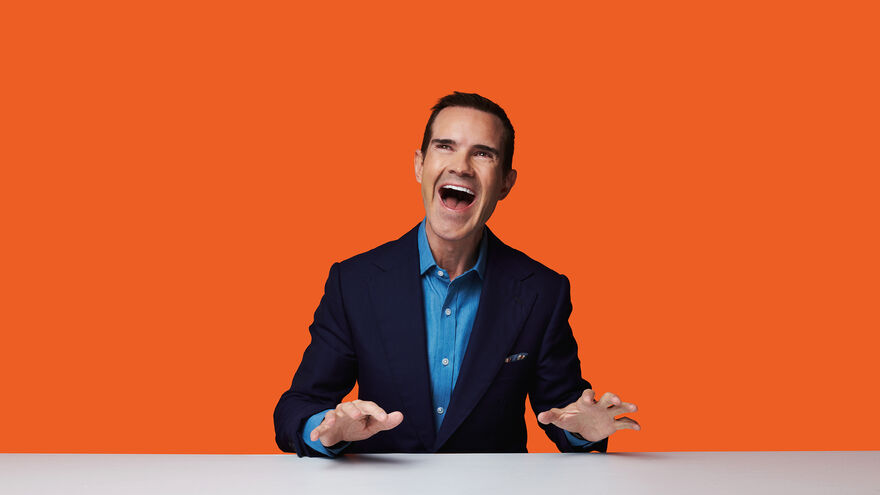 Jimmy Carr, a white man with short black hair is sat at a white desk. He is wearing a light blue shirt and a navy blue jacket. He is laughing and has both his hands slightly raised from the desk.