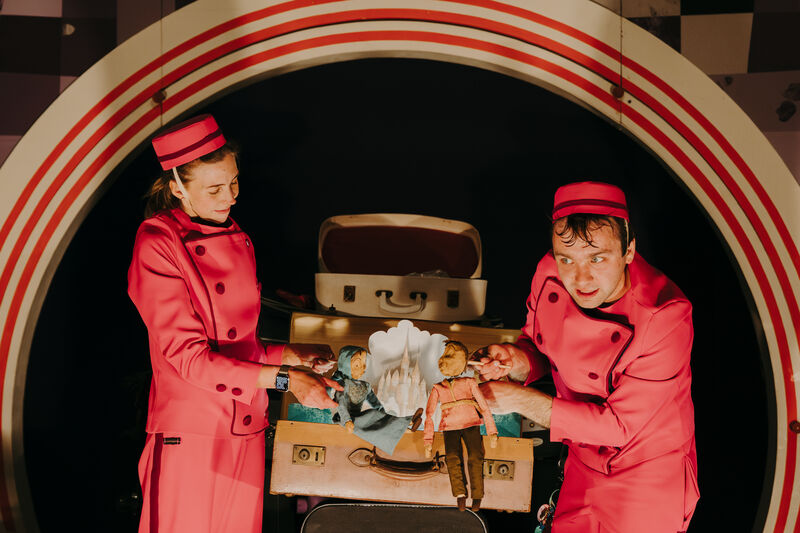 A man and a woman in red jumpsuits and hats are holding up two dolls in front of an open vintage suitcase. There is an arched backdrop behind them. 