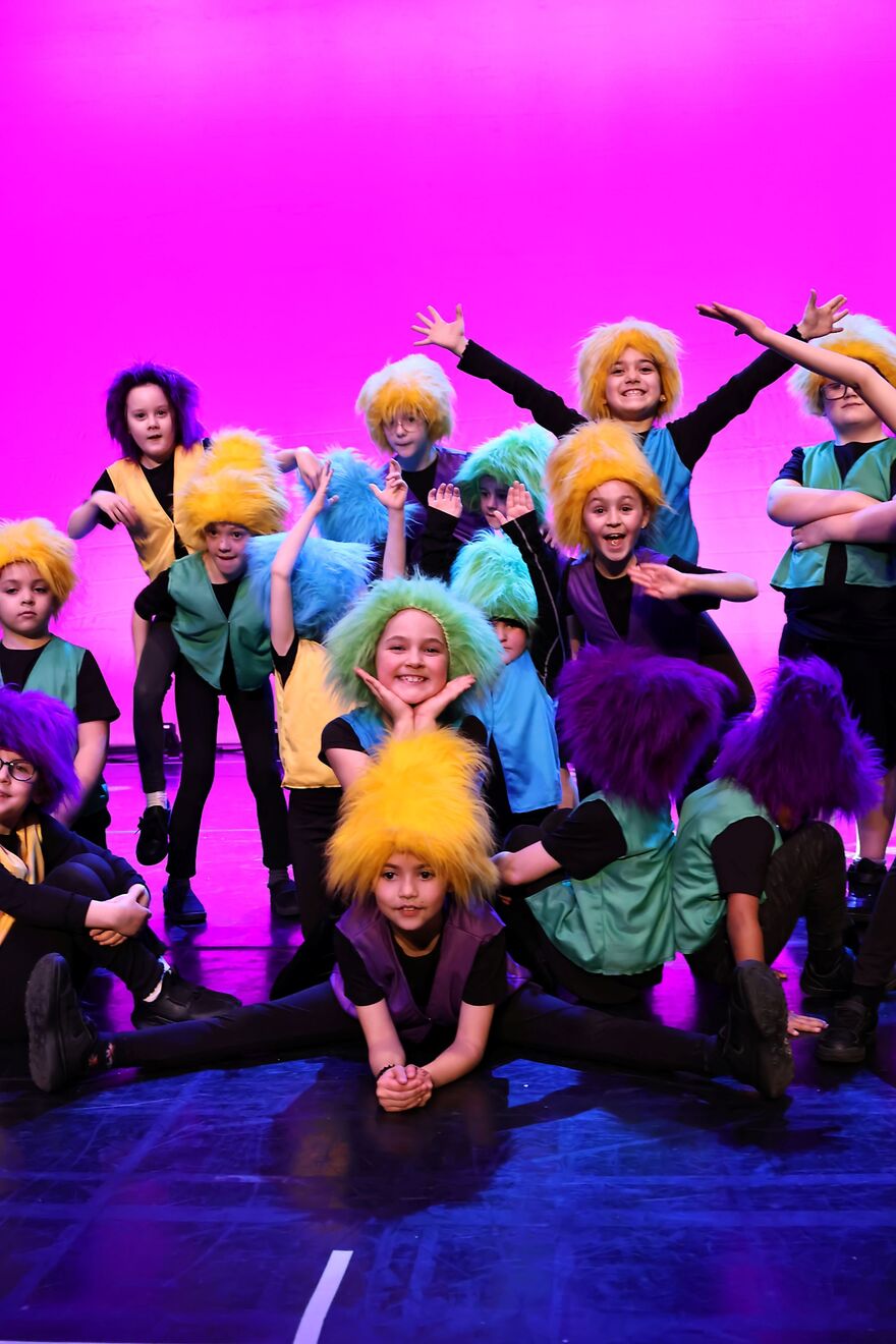 A group of young dancers on a stage wearing fuzzy yellow, purple and green wigs. They are in various positions, including the splits, holding arms up and waving