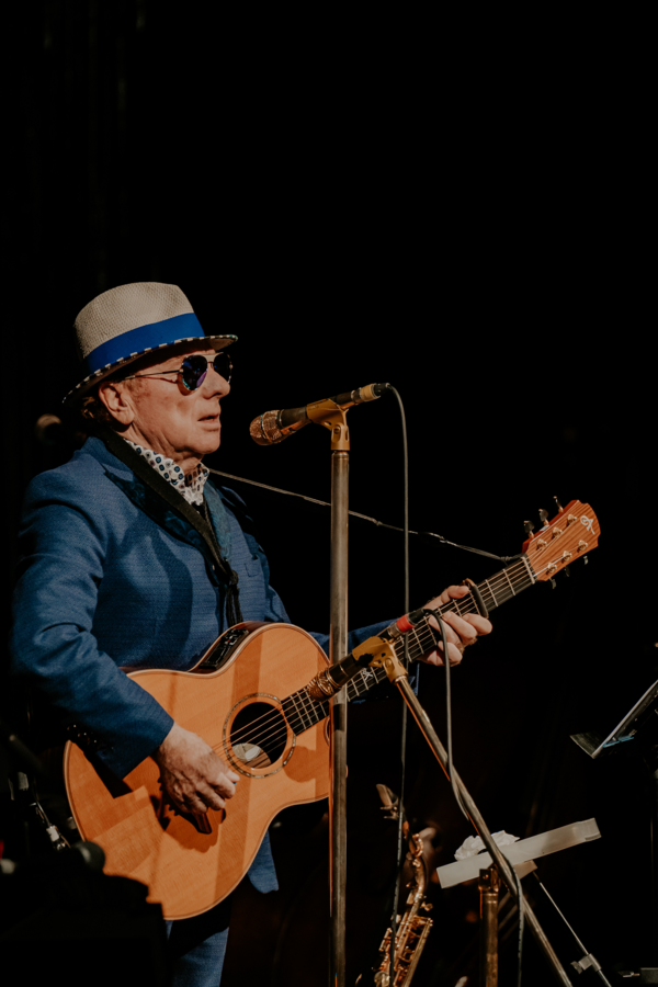 Van Morrison performing on stage. He's a white man in his 70s wearing a suit, hat and sunglasses, holding an acoustic guitar and singing into a mic