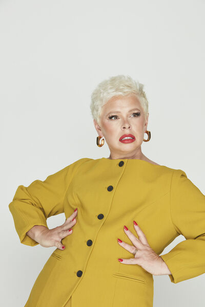 A woman with short blonde hair wearing gold hoops and red lipstick. She has her hands on her hip and wearing a yellow blouse