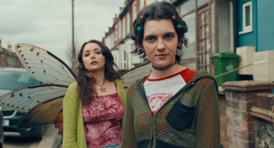 Two young women look sternly into the camera. They are dressed in 90s clothing and one of them wears a pair of fairy wings. They are stood on an urban street 