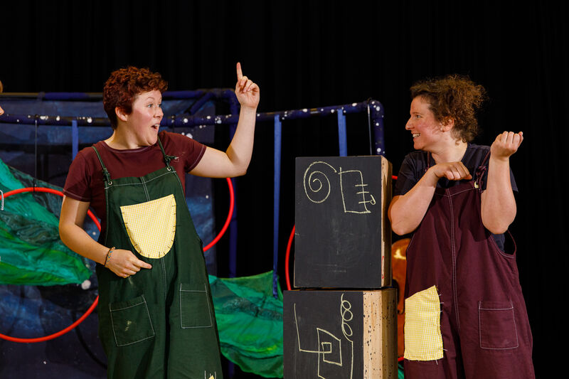 Two people wearing patchwork dungarees standing with one miming playing the violin and the other pointing upwards