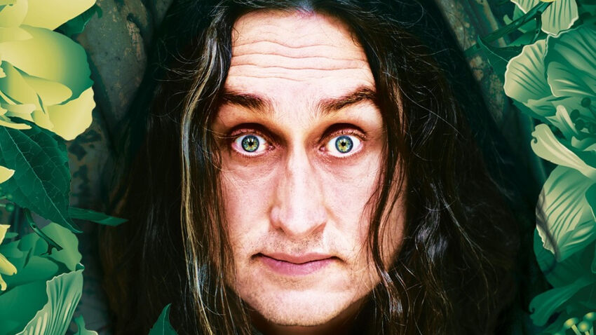 Man with long hair looks directly forwards in a leafy background with a slightly surprised expression