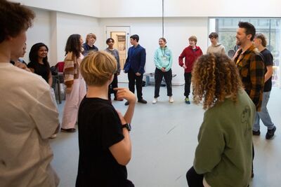 Young comedians stand in a circle in a bright room, the comedy workshop leader stands to the right