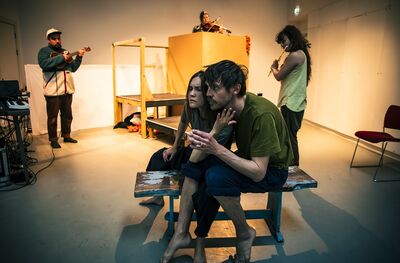 In a dimly lit room two people sit closely together on a bench whilst behind them three musicians play instruments