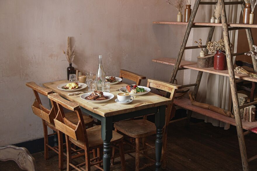A wooden table with four chairs around it, there a plates filled with food and empty glasses on the table. To the side of the table is a wooden a-frame bookshelf