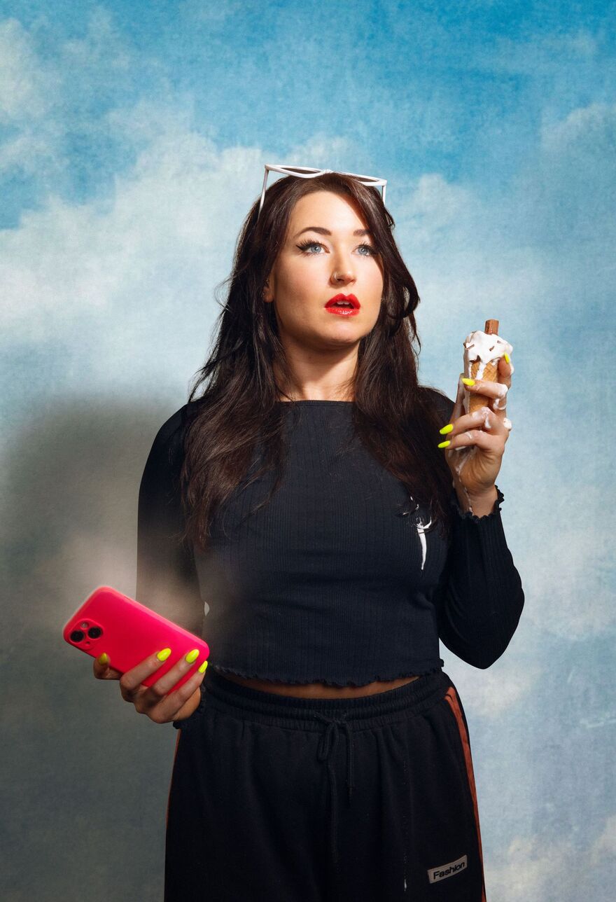 A woman with brown hair wearing white sunglasses on  her head and a black outfit. She is holding a bright phone in her left hand and a melting ice cream in the other