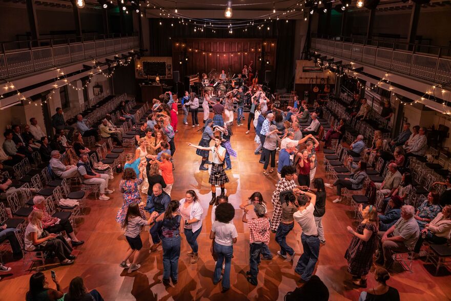 A caller stands in the middle of audience members as they dance in a hall