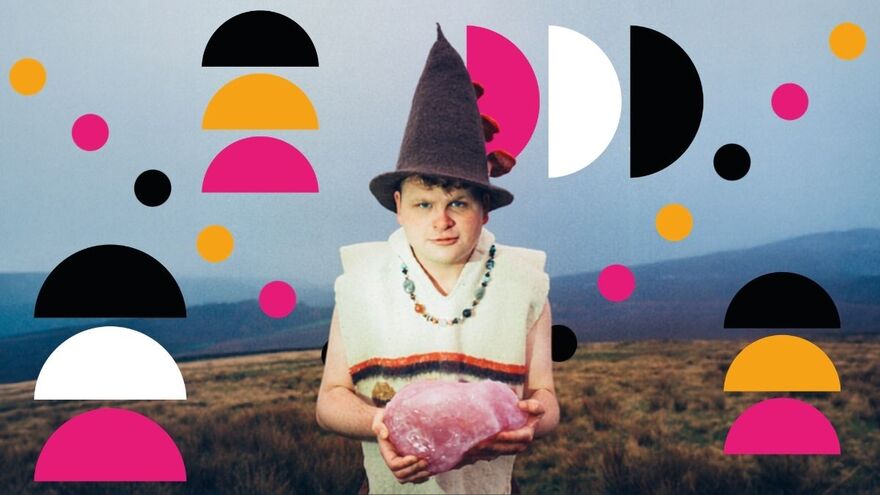 A man stands on the moors wearing a pointy wizard hat and white vest holding a large pink quartz with both hands