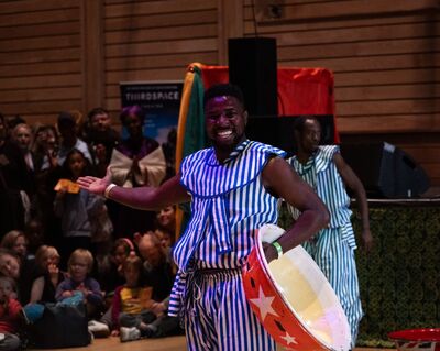 A person smiling wearing a blue stripey outfit. He is holding a drum with his left hand and holding his other arm out.