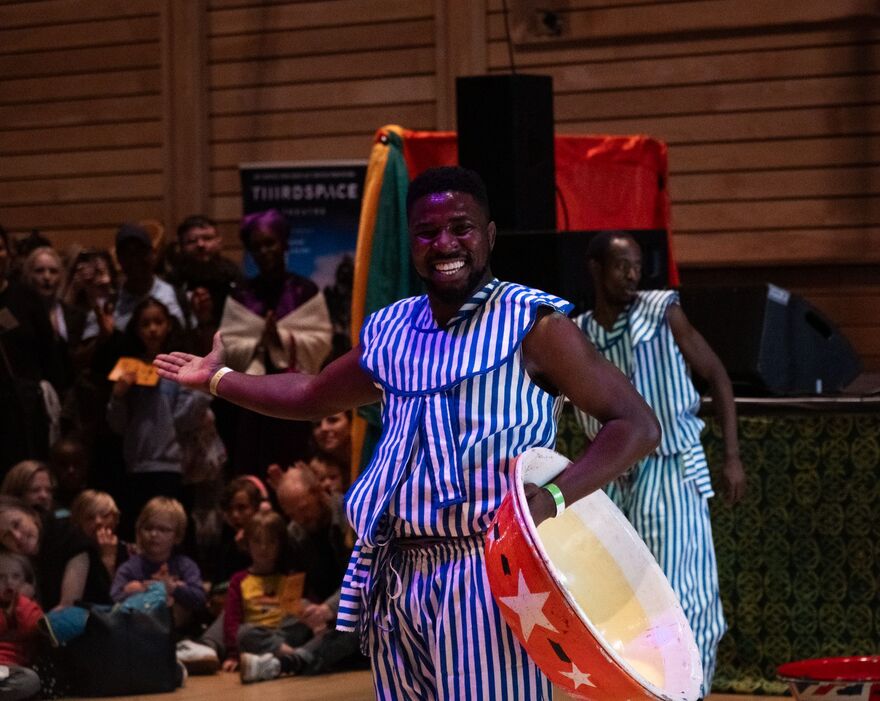 A person smiling wearing a blue stripey outfit. He is holding a drum with his left hand and holding his other arm out.