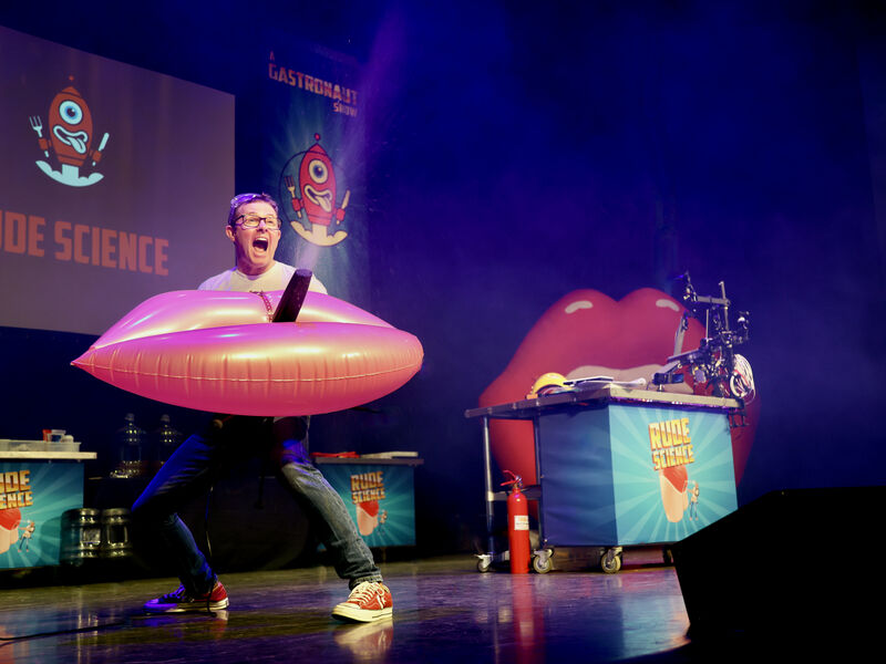 Man stands on stage and holds inflatable pair of lips that's spouting water through the centre