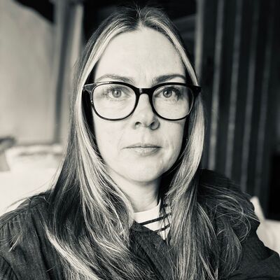 A white woman with long blonde hair, wearing large dark rimmed glasses, a stripy t-shirt and dark over jacket