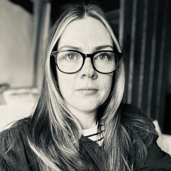 A white woman with long blonde hair, wearing large dark rimmed glasses, a stripy t-shirt and dark over jacket