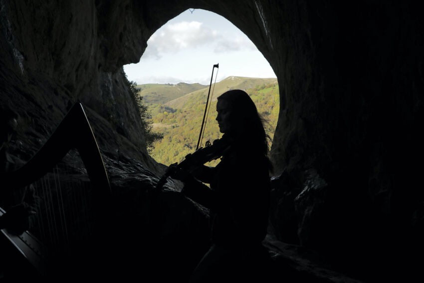 A violinist and harpist performing in a cave, silhouetted against a background of green hills in the distance