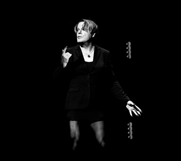 Eddie Izzard in a black blazer and dress wearing fishnet tights and a pendant necklace