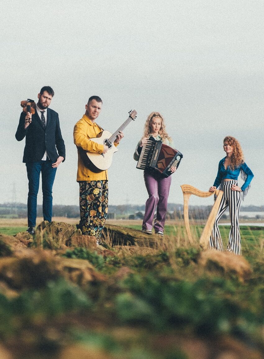 The 4 members are stood in a field, front-facing. From the left, the tall, brown-haired male violinist rests his violin on his left shoulder and wears a blue suit with blue jeans and a tie. The dark-haired male guitarist holds his acoustic guitar under his left arm and atop his bright yellow jacket, wearing floral trousers. The blonde-haired female accordionist wears her hair tied in a wavy ponytail and holds her accordion over her torso, wearing purple trousers. Lastly, the female harpist leans her arm over the top of the harp and wears a blue-fringe top with black-and-white striped trousers.