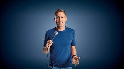 Russell Howard wearing a blue top and holding a microphone, he is standing in front of a blue background