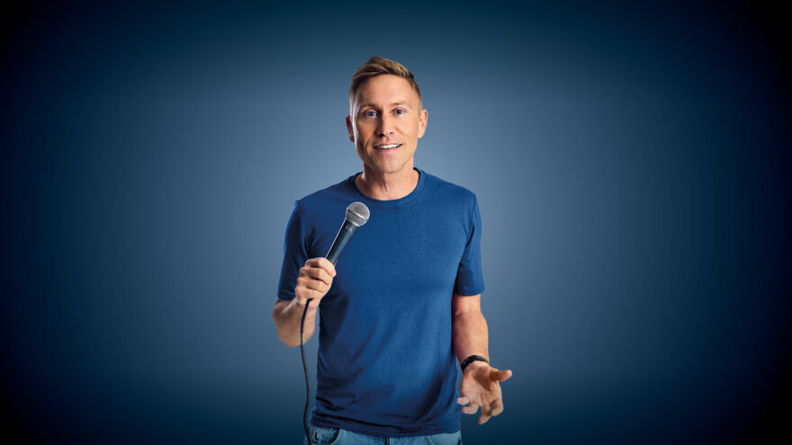 Russell Howard wearing a blue top and holding a microphone, he is standing in front of a blue background