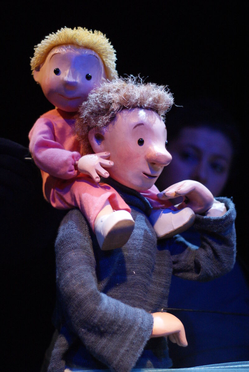A wooden puppet of a man walking and carrying another wooden puppet of a toddler on his shoulders