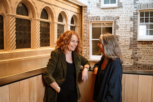 Two women looking at each other smiling. They are wearing black tops and dark coloured blazers. They are standing in front of a brick wall with arched windows