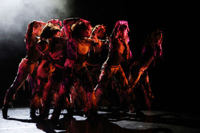 A group of dancers on stage with their arms up around their faces. There is a red spotlight on them and smoke around them