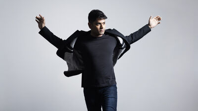 Conductor Alpesh Chauhan stands against a light gray background with his arms outstretched, looking to his right as the jacket flares slightly around him.