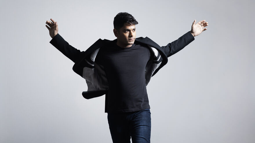 Conductor Alpesh Chauhan stands against a light gray background with his arms outstretched, looking to his right as the jacket flares slightly around him.