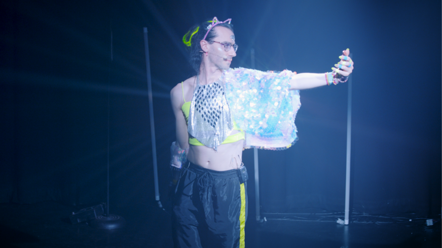 A non-binary person wearing neon clubbing attire stands holding one arm out, looking at a phone.