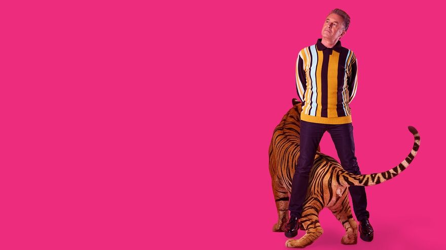 Chris Packham standing with his arms behind his back and his legs apart. He has a tiger standing in between his legs facing away. They are standing over a bright pink background.