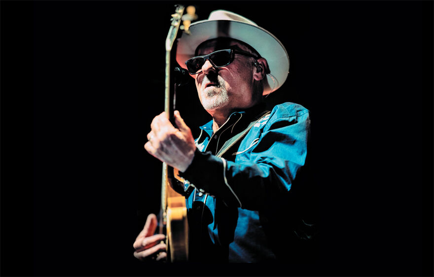 A white man playing guitar. He wears a white hat and blue denim shirt.