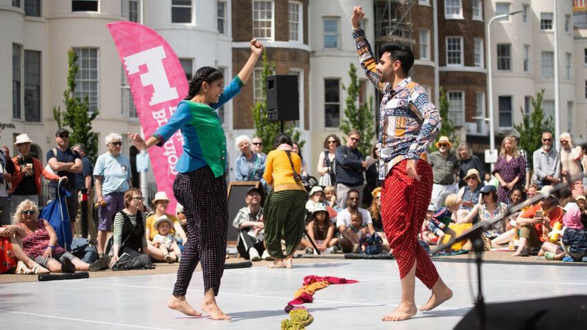 Two performers are stood facing each other with their arms in the air. They are dressed in colourful outfits and are surrounded by the audience.