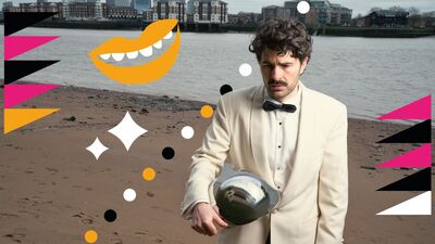 Paddy Young standing on a beach wearing a cream suit and black bowtie looking at the floor holding a cloche under his arm