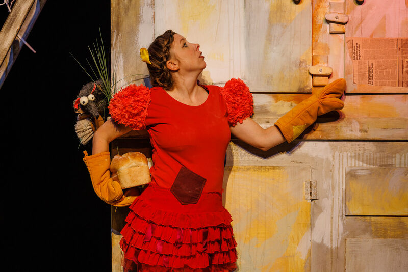 A woman dressed as a red hen stands in the shape of a teapot wearing oven gloves and holding a load of bread