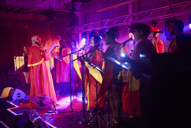 Fatt Butcher stands facing and directing a choir on stage, with purple mood lighting illuminating their red and yellow robes