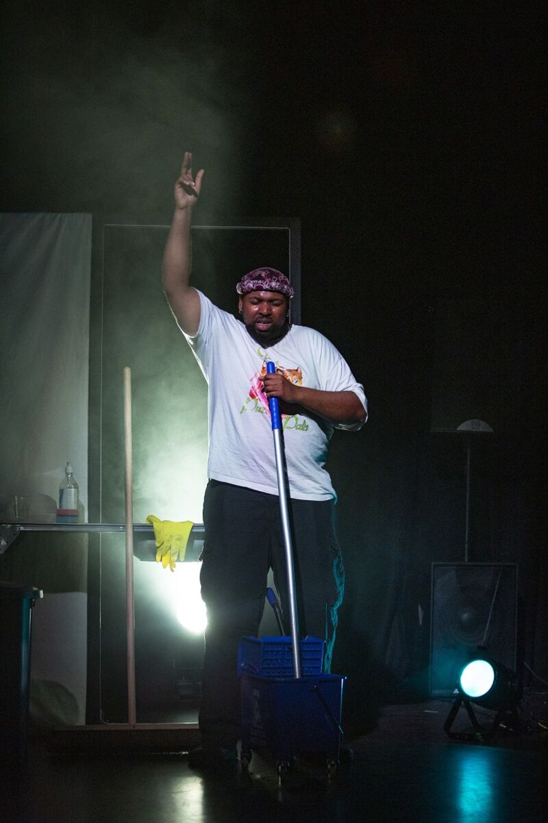 A black man in a white t-shirt and black trousers stands in front of a mop bucket, whilst holding on to the mop handle with his left hand. His right arm is raised, fingers pointing towards the ceiling.