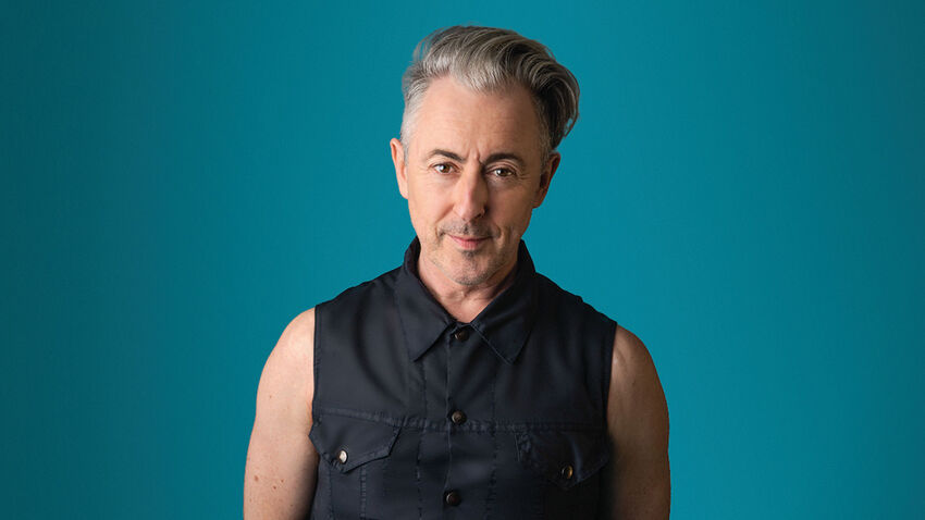 Alan Cumming wearing a dark blue sleeveless shirt with a teal colour background